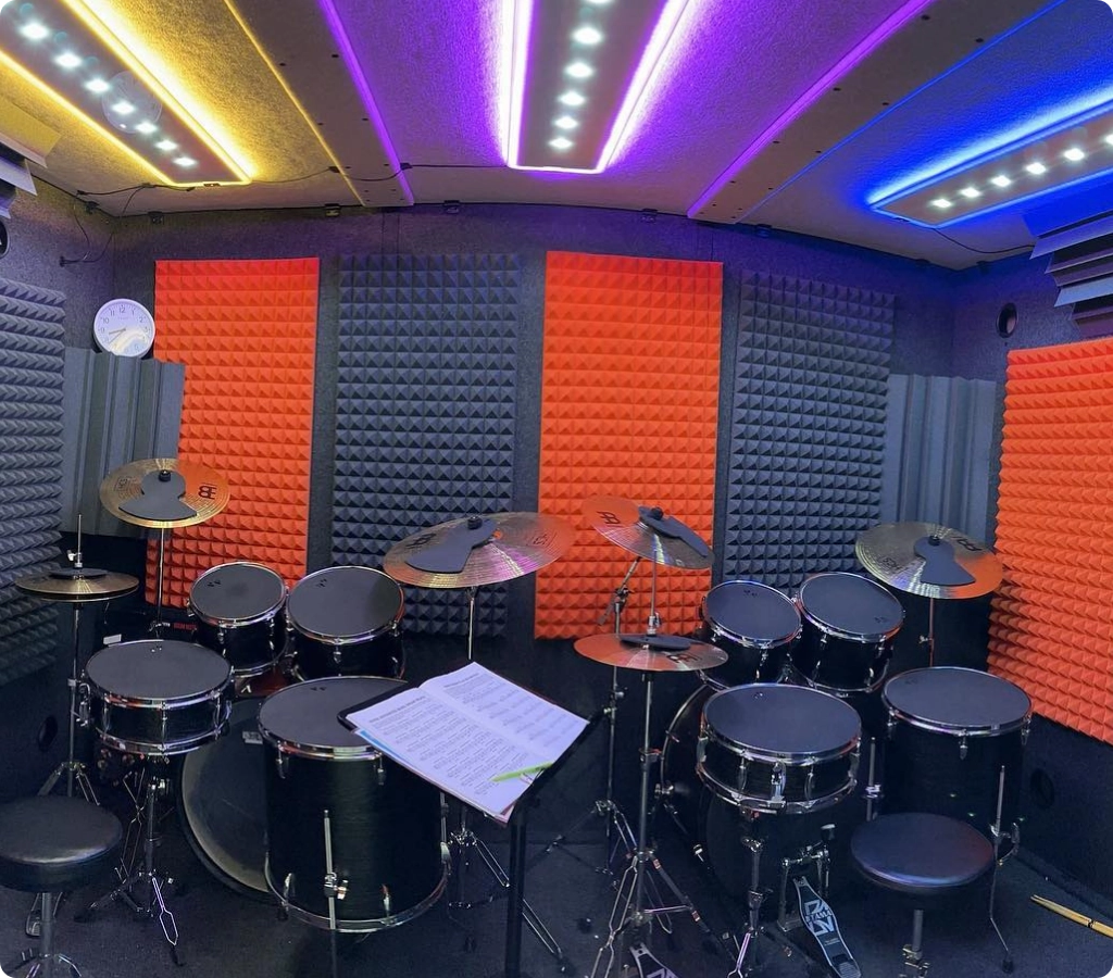 Large acoustic drum kit inside a WhisperRoom booth with orange and gray acoustic treatment panels and overhead lighting.