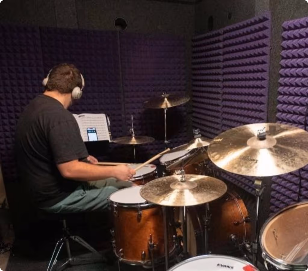 Drummer practicing inside a WhisperRoom booth with purple acoustic foam, demonstrating real isolation compared to plexiglass drum shields.