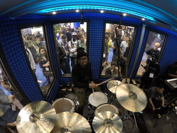 Drummer demonstrating gear inside a WhisperRoom sound isolation booth at the NAMM Show, with attendees watching from the noisy show floor outside