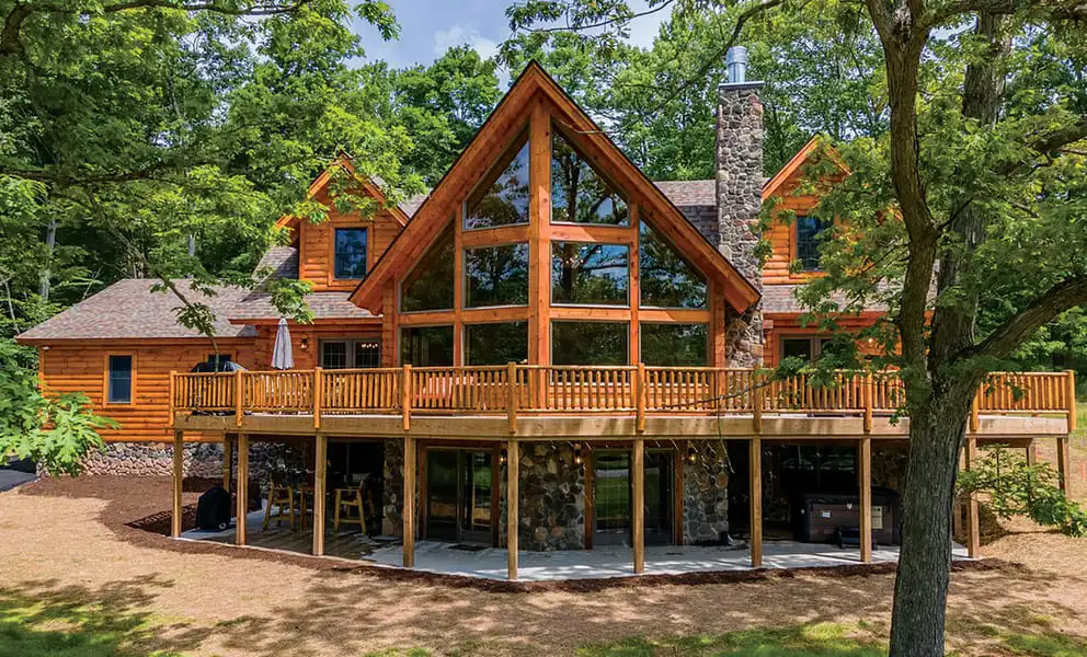 Exterior view of the Clear Fork log home by Hochstetler Log Homes, showcasing a majestic timber design with a wall of windows, stone fireplace chimney, and wraparound deck surrounded by trees.