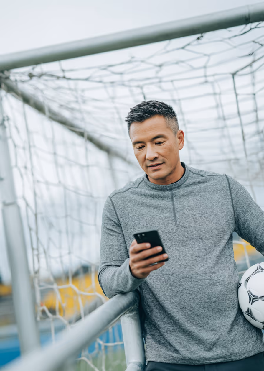 Man in gray shirt leaning on soccer goalpost holding ball and looking at his phone.