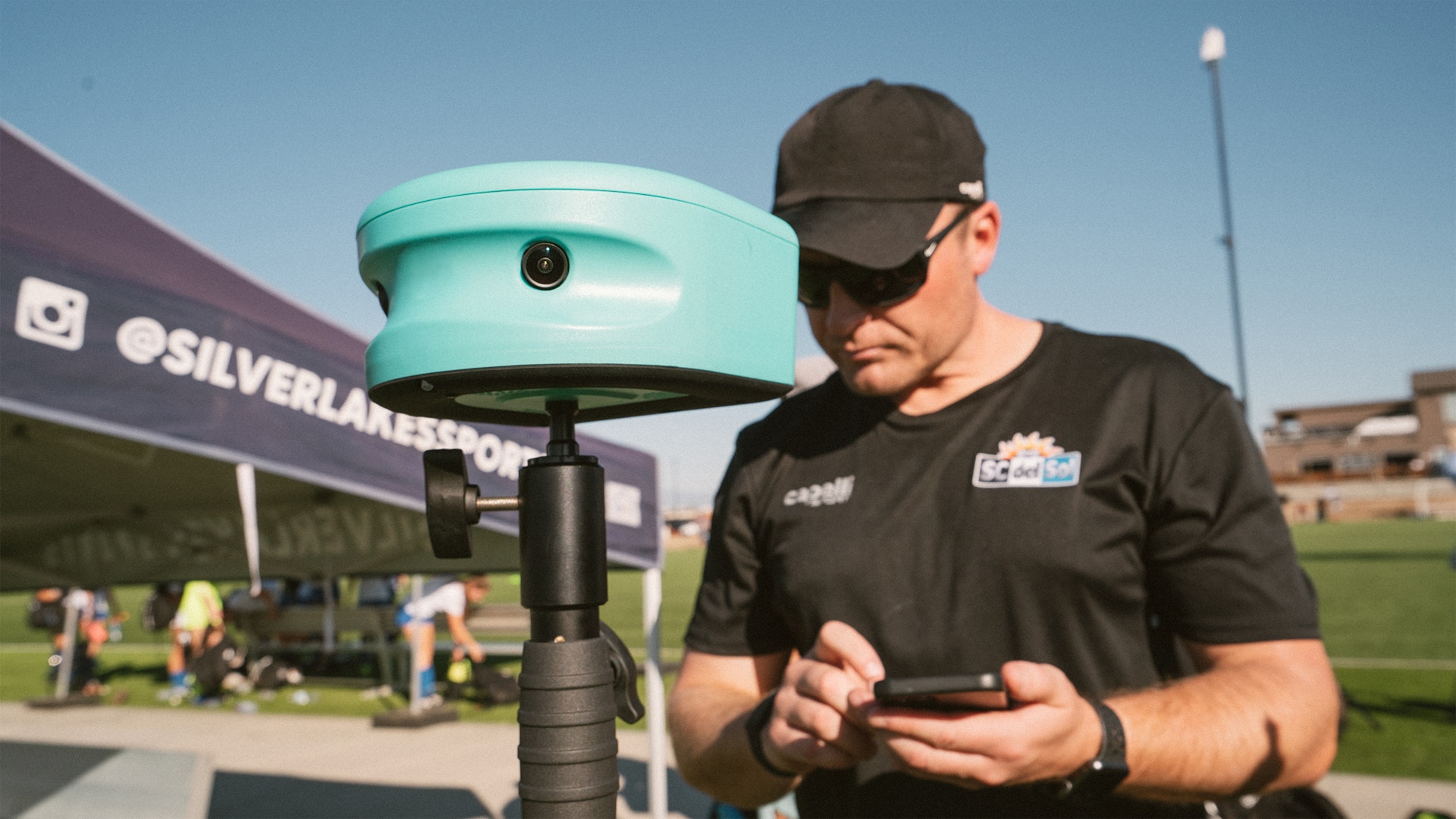 Adult setting up a Trace AI soccer camera on a tripod at the sideline of a youth soccer field while using the Trace app on a phone