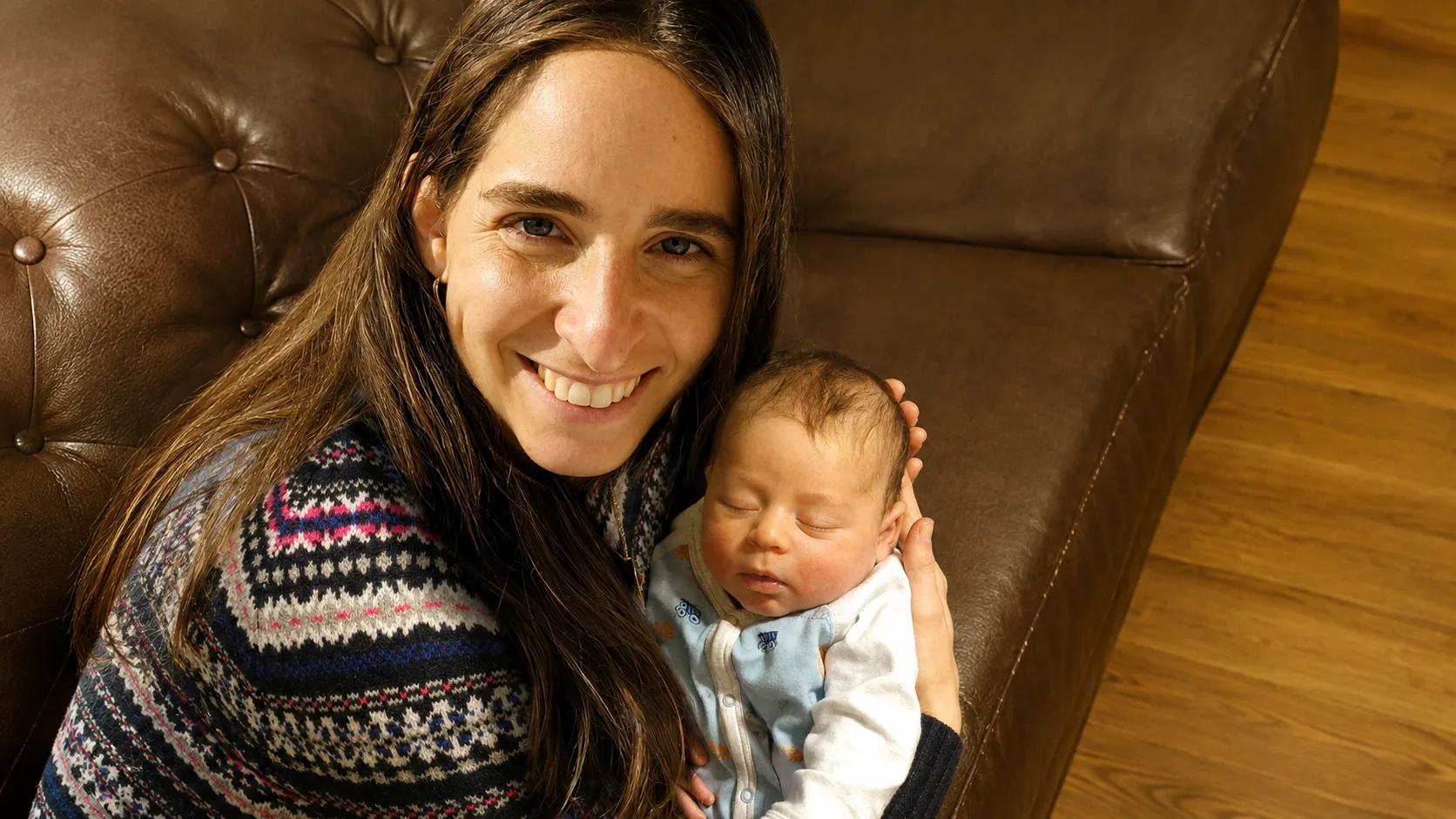 Smiling woman holding a baby who looks up at her, indoor setting with light and dark walls.