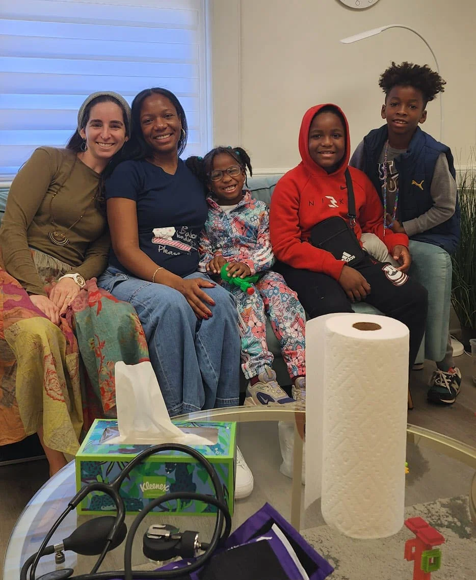Two smiling women and three children sitting together on a sofa, with a blood pressure cuff and stethoscope on the table in front of them