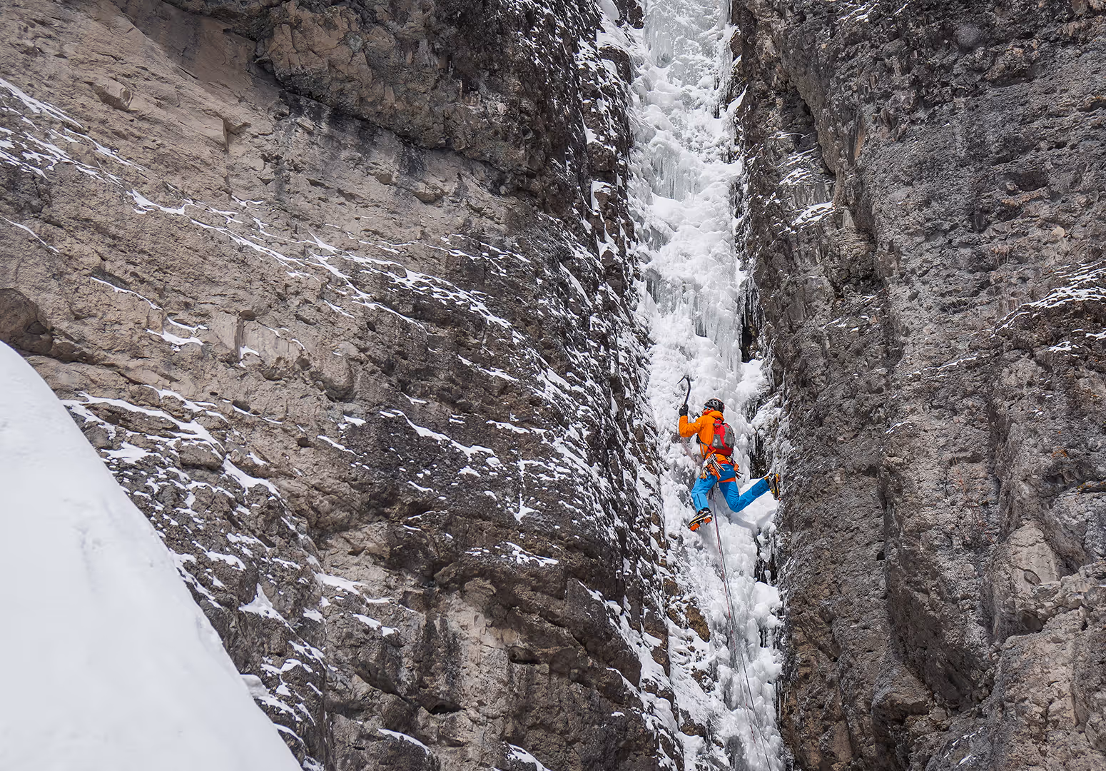 A person wearing bright winter gear is climbing a steep, narrow ice formation on a rocky cliff.