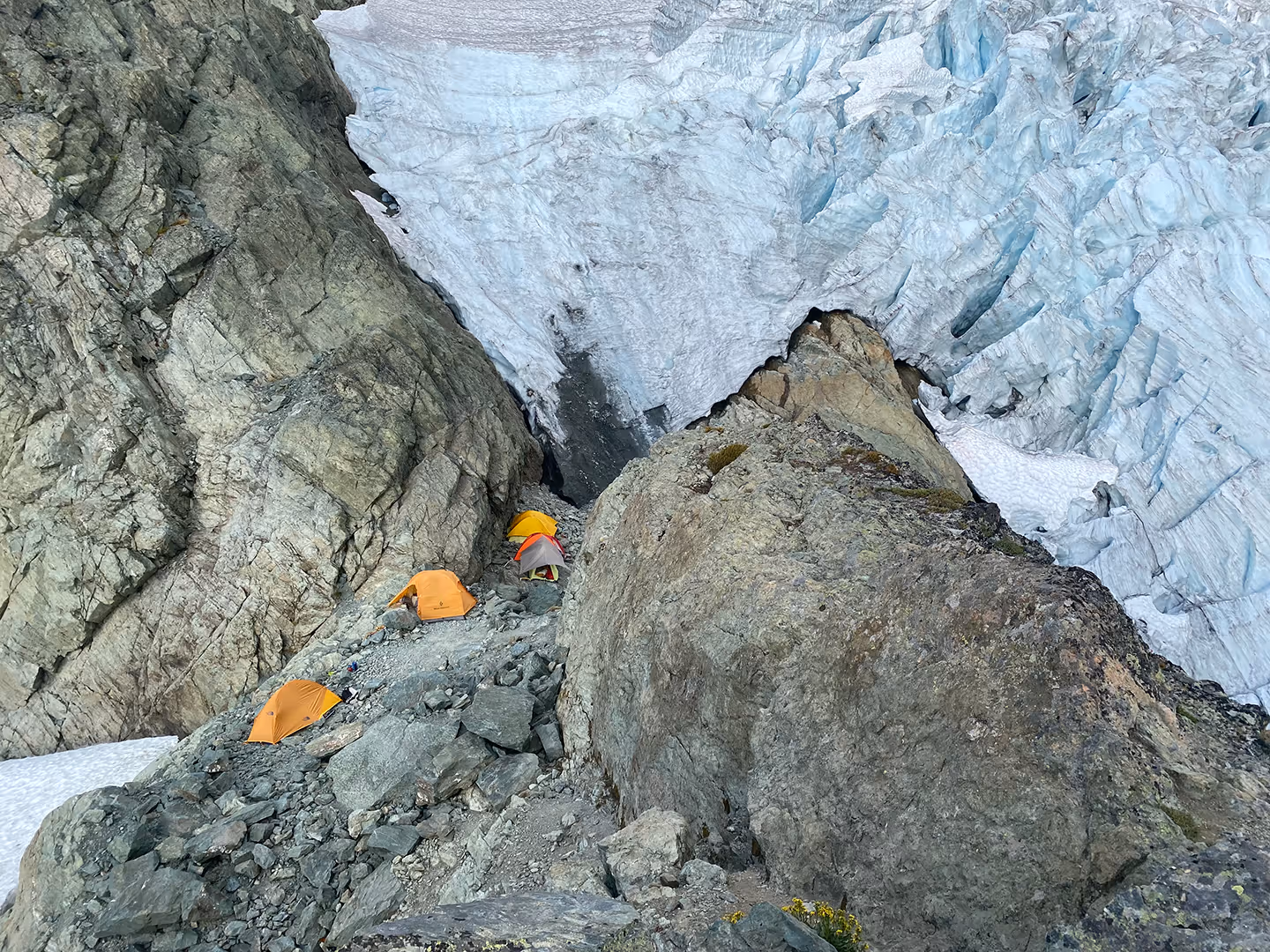 Three orange tents set up on rocky terrain next to a large glacier.