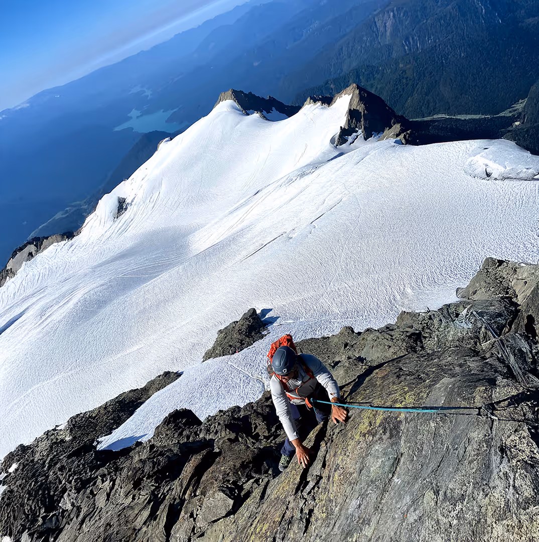 A climber wearing a helmet ascends a rocky slope with a snowy mountain landscape in the background.