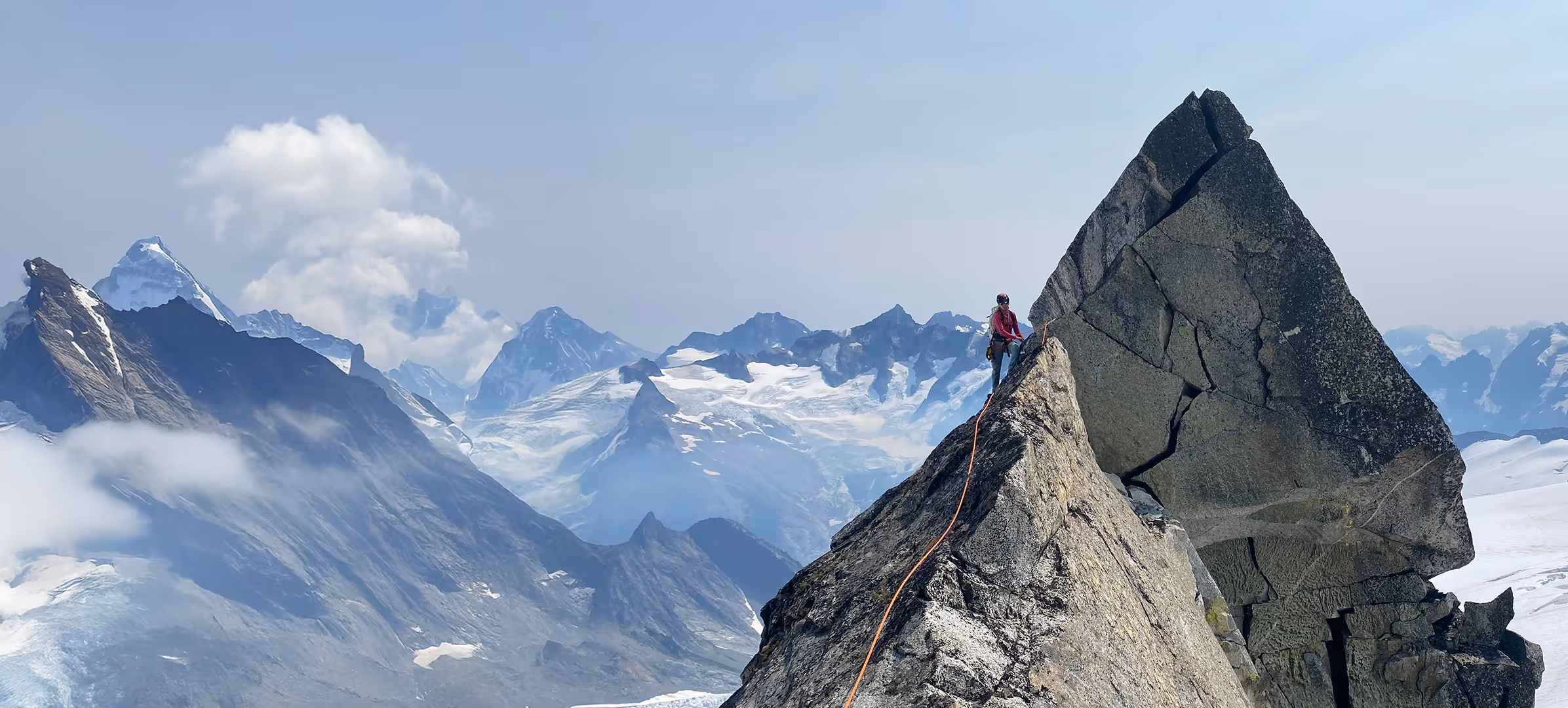 Person climbing a steep, rocky mountain peak with snow-covered mountains in the background.