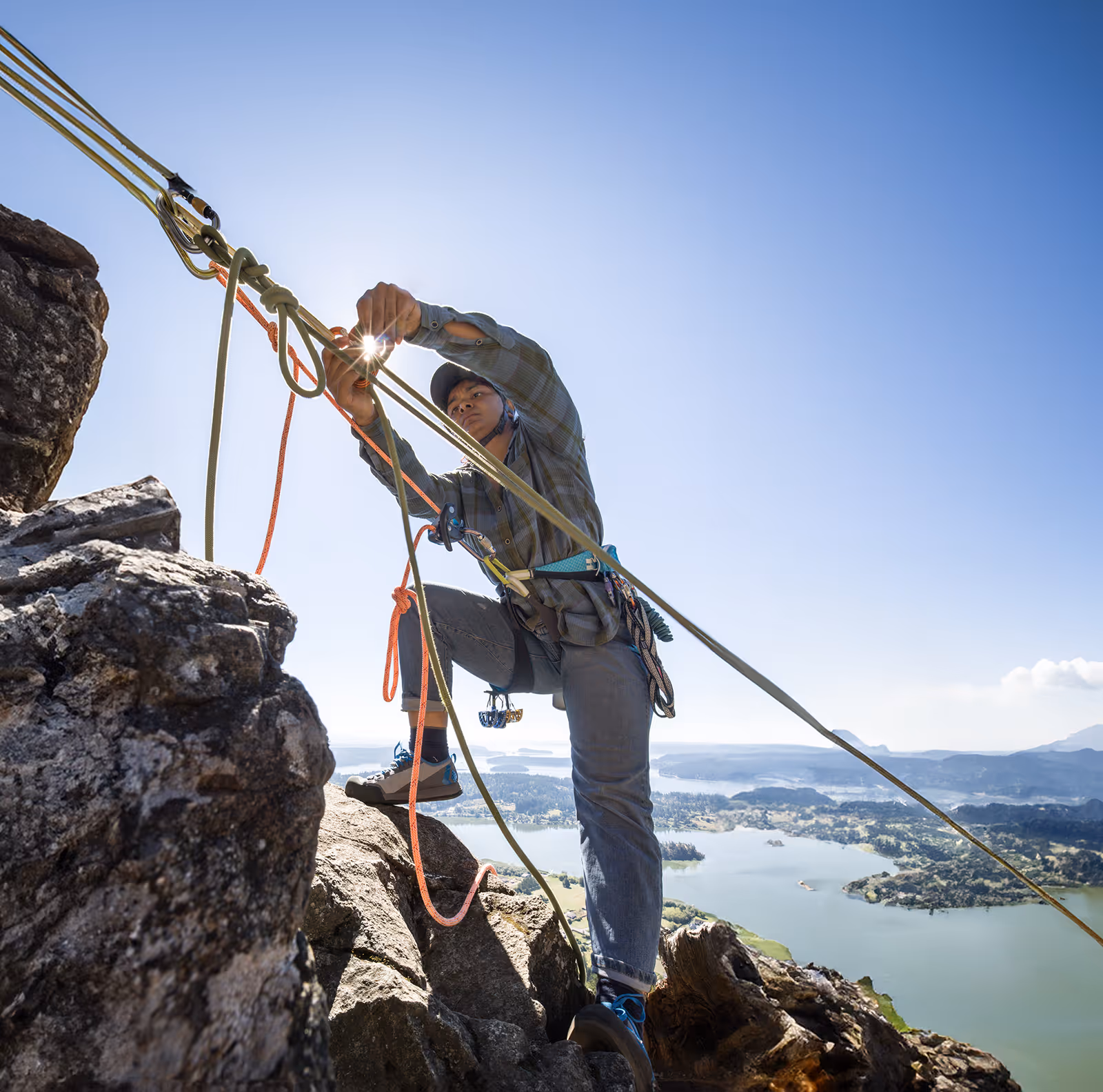 A person in climbing gear adjusts ropes on a rock, overlooking a lake and mountains.