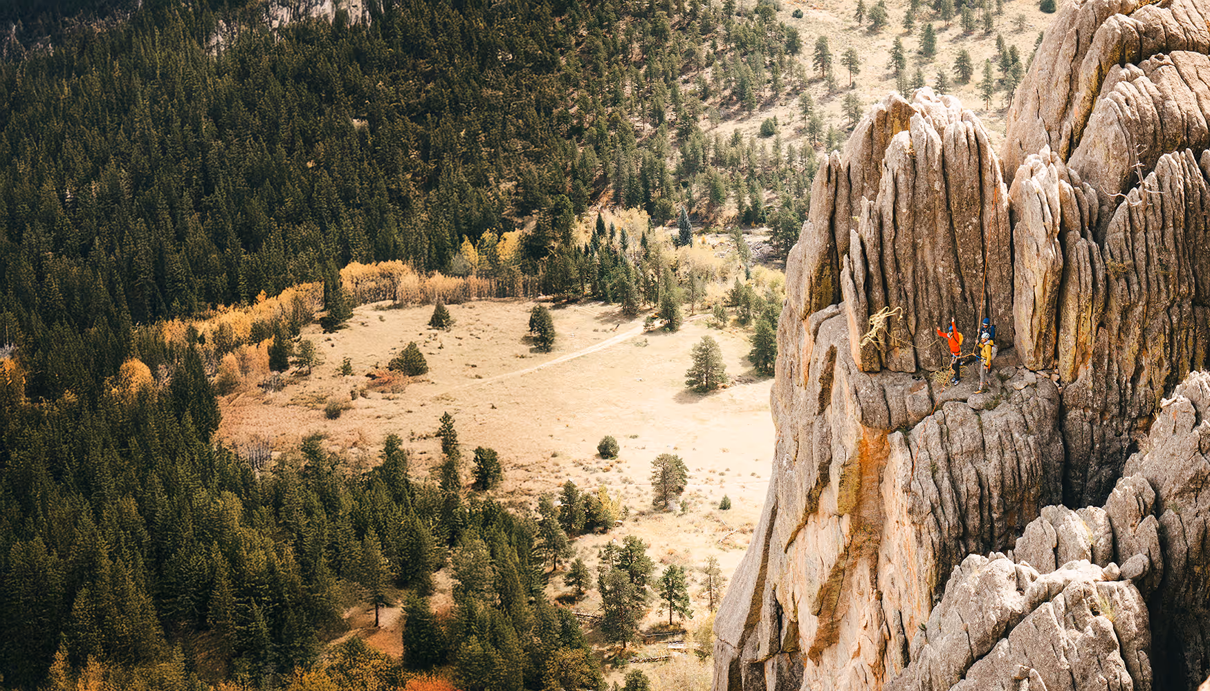 Two people in climbing gear stand on a large rock formation, overlooking a forested area.
