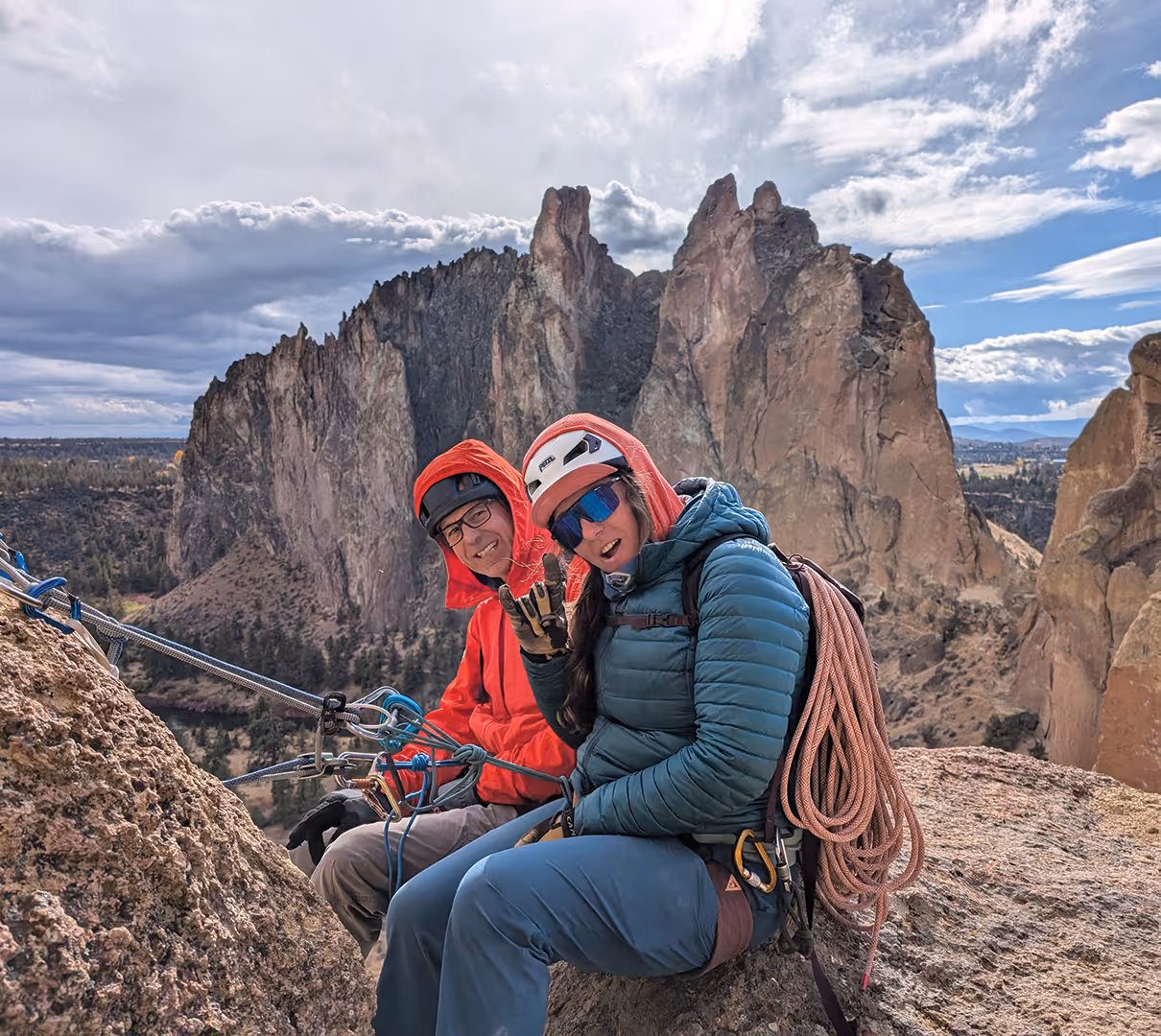 Two climbers in helmets and gear smile while resting on a rock with a mountain in the background.