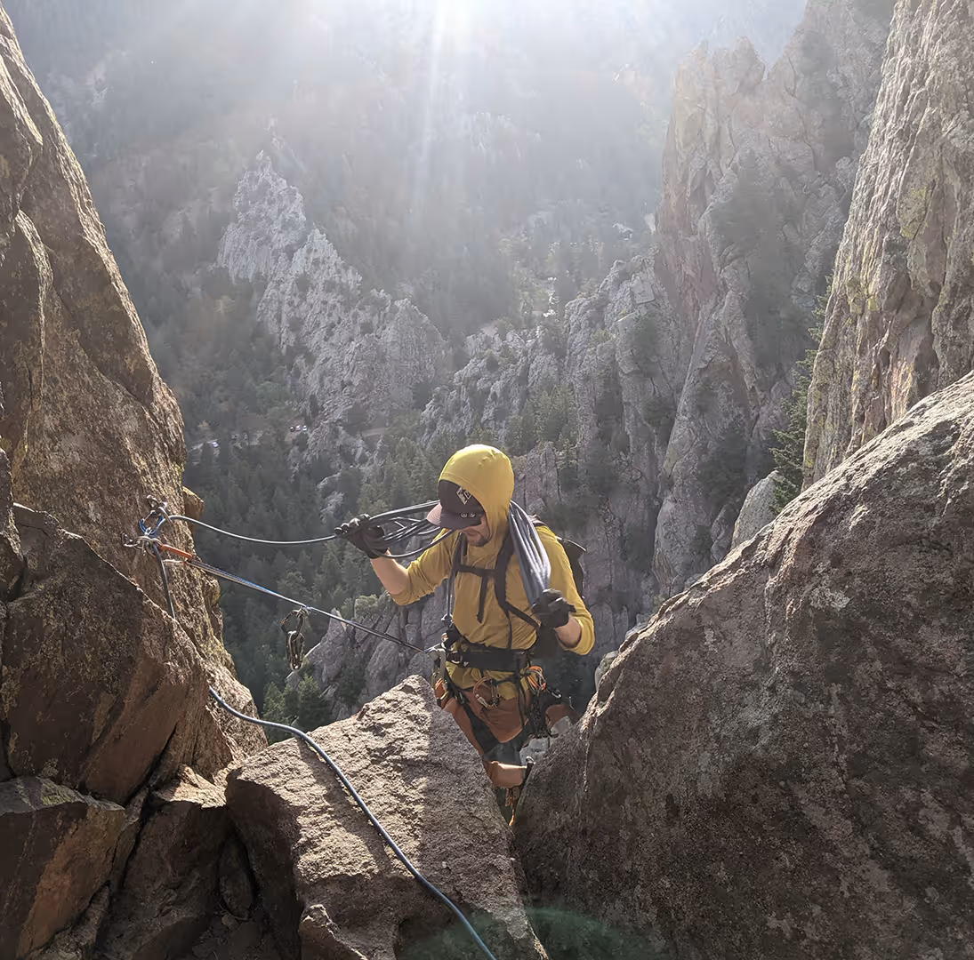 A climber in a yellow jacket ascends a rocky mountain using ropes on a sunny day.
