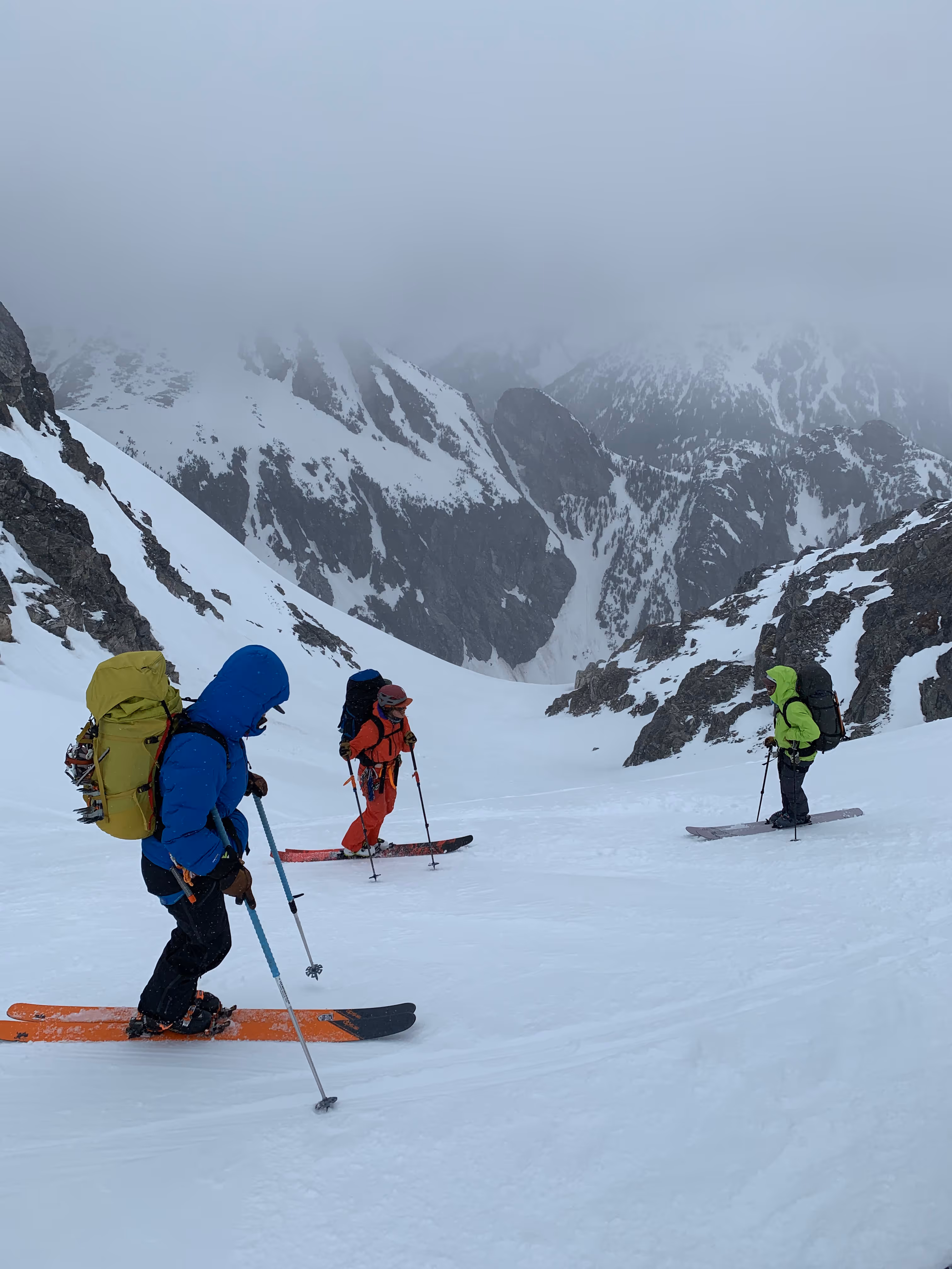 Three people skiing downhill on a snowy mountain in foggy weather, wearing colorful gear and backpacks.