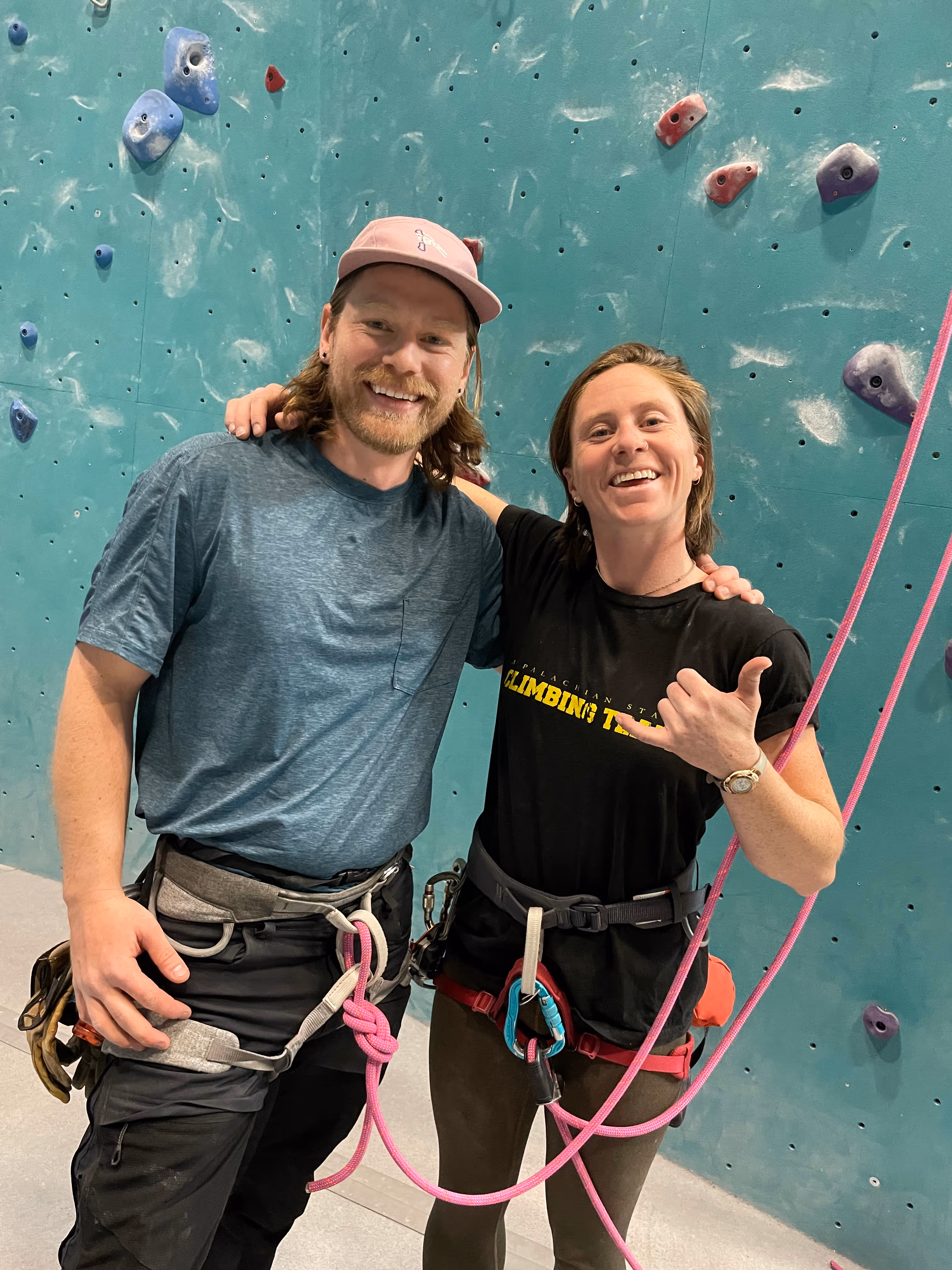 Two people wearing climbing gear smile and pose in front of an indoor climbing wall.