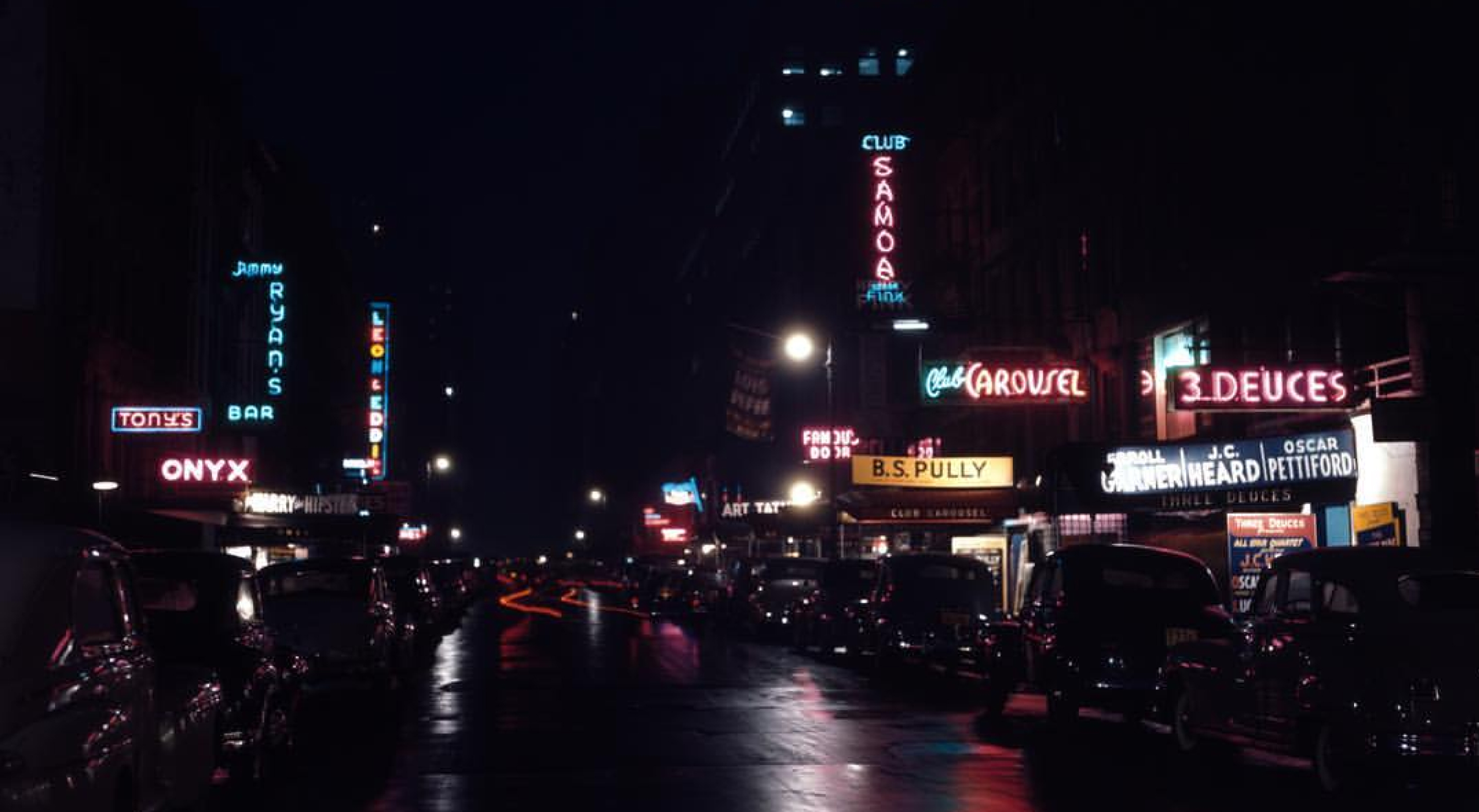 Night street scene with vintage cars parked along both sides and illuminated neon signs for bars and clubs.