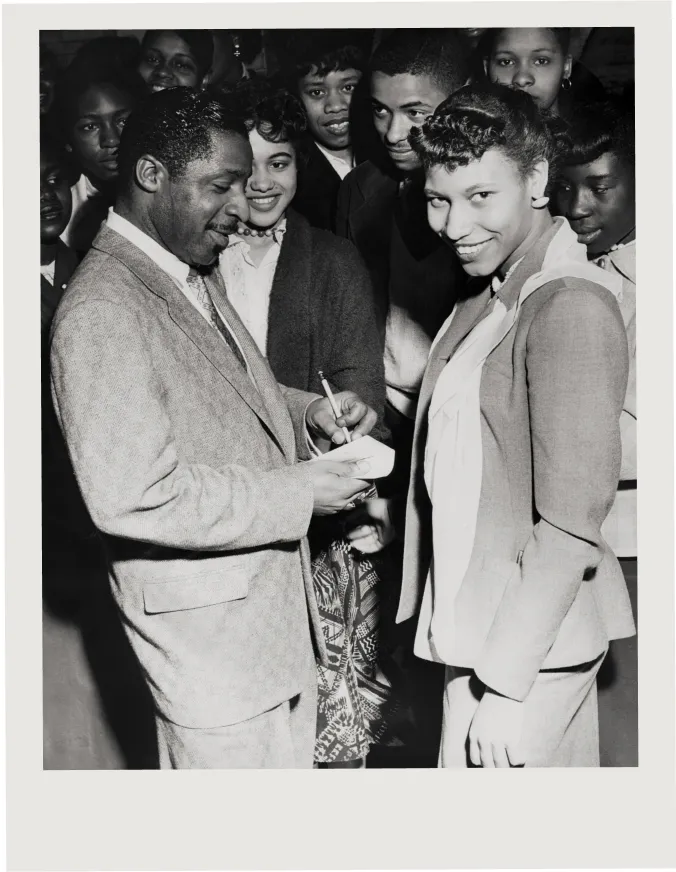 Erroll Garner signing an autograph for a smiling woman while a group of people watches in the background.