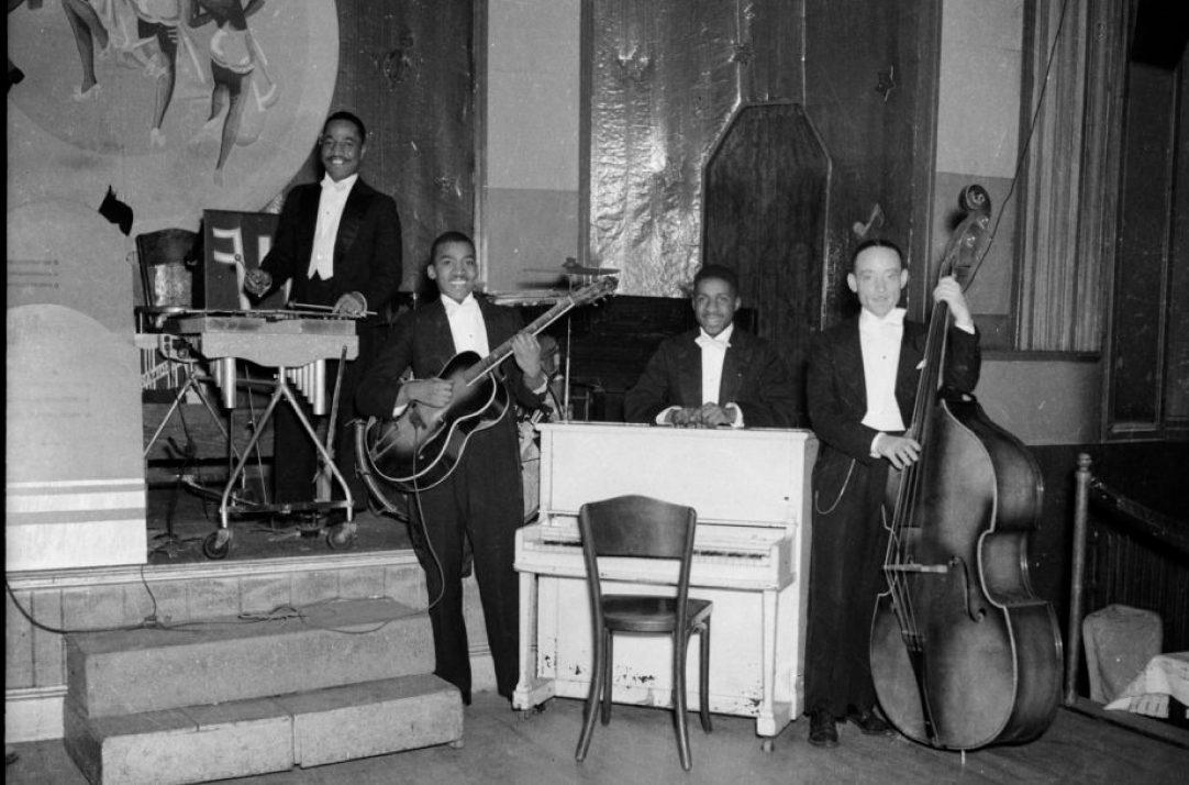Black and white photo of four male musicians in tuxedos posing with vibraphone, guitar, piano, and double bass in a performance venue.