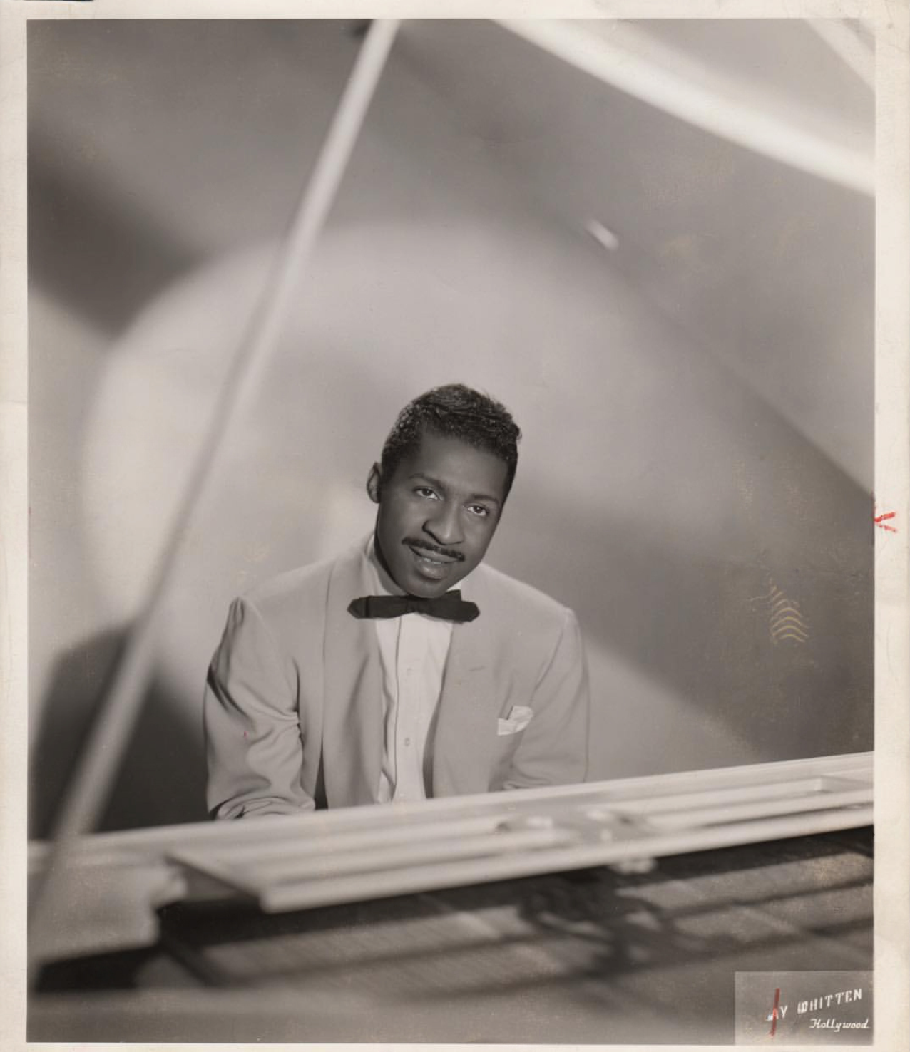 Black and white portrait of Erroll Garner in a light-colored suit and bow tie sitting at a piano.