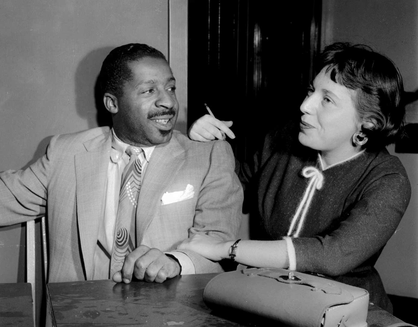 Black and white photo of Erroll Garner in a suit with patterned tie smiling at a woman who is holding a cigarette, seated at a table with a handbag.