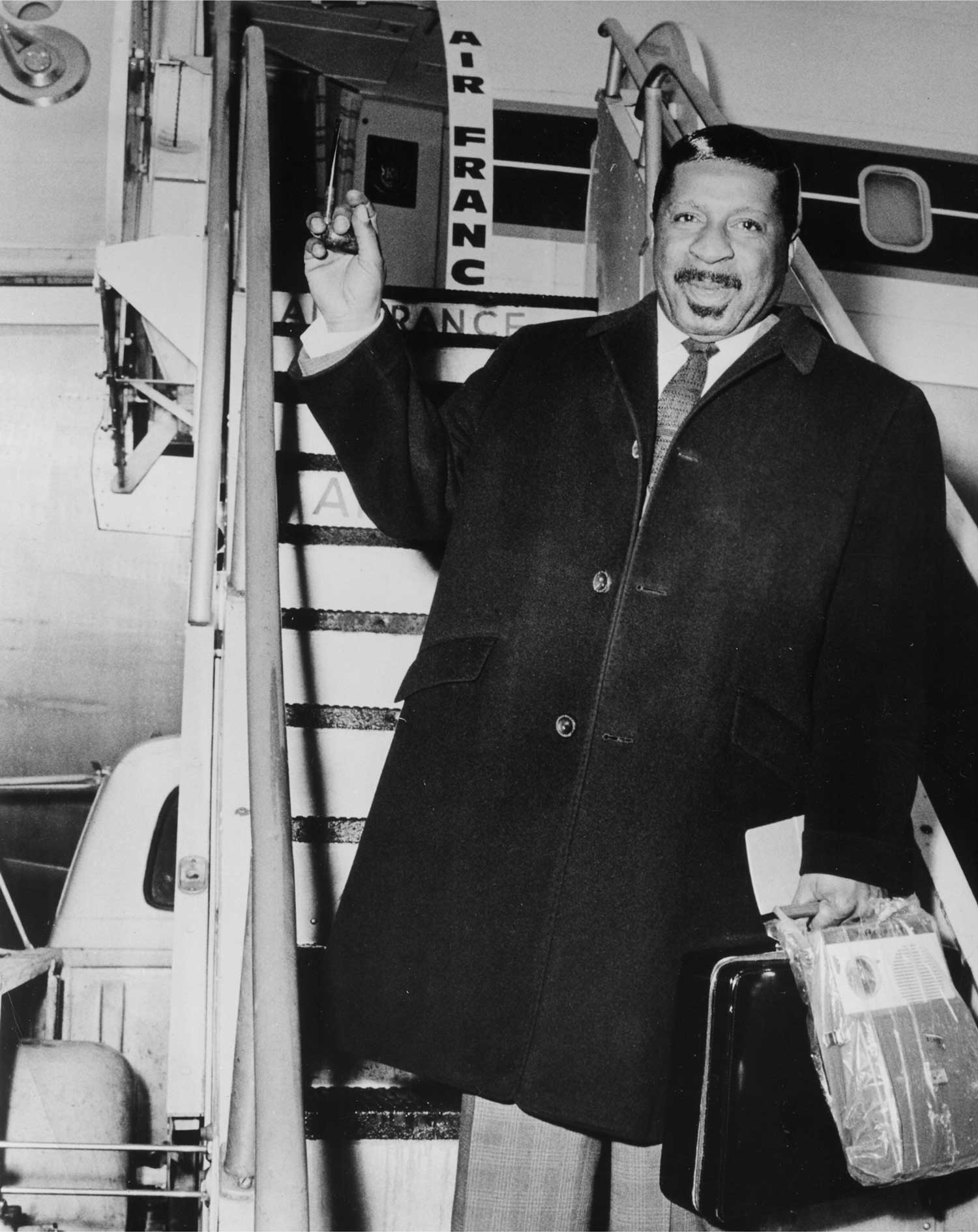 Erroll Garner in a dark coat standing at the base of airplane stairs holding a briefcase and a plastic-wrapped package.