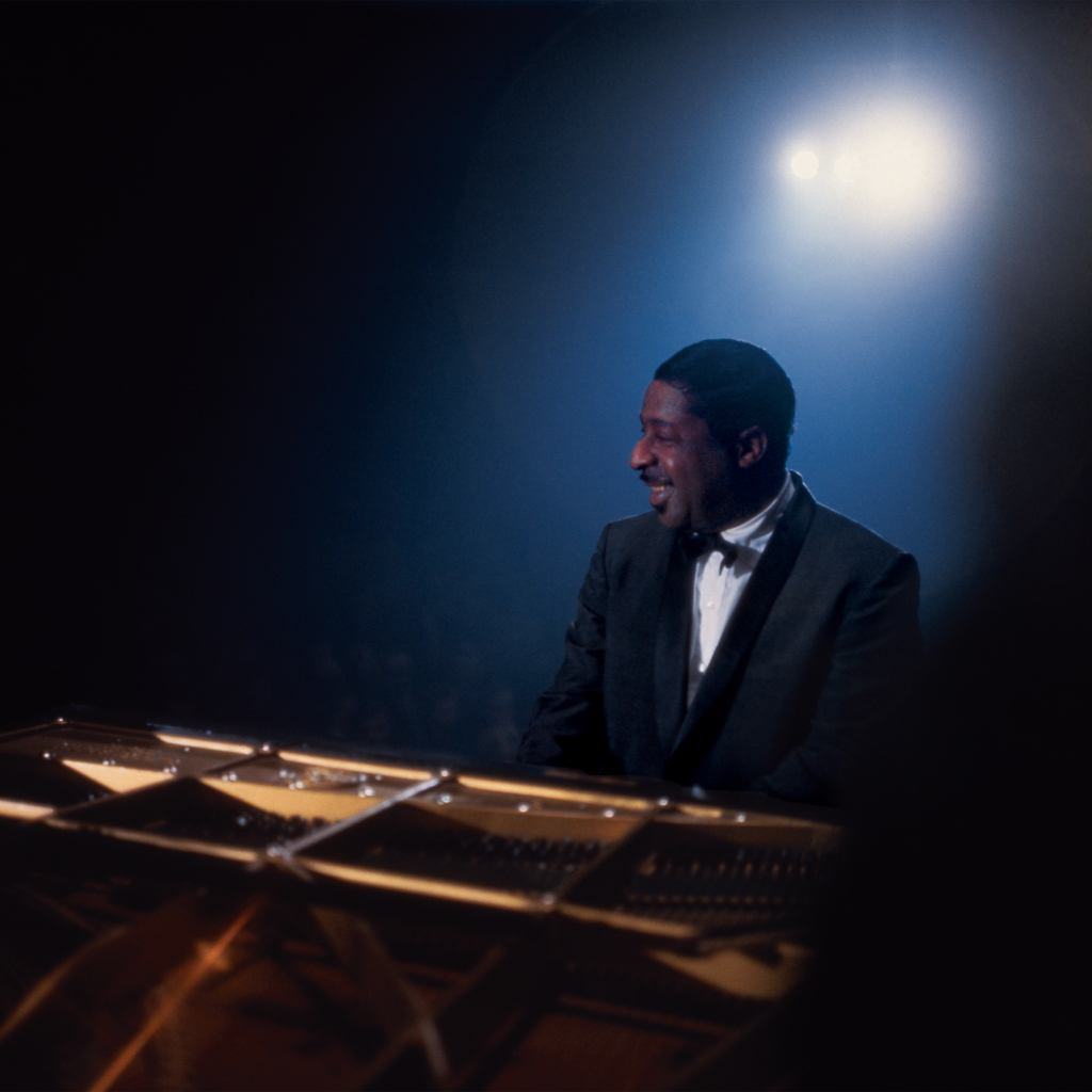 Erroll Garner in a tuxedo smiling and playing a grand piano on a dimly lit stage with a spotlight behind him.