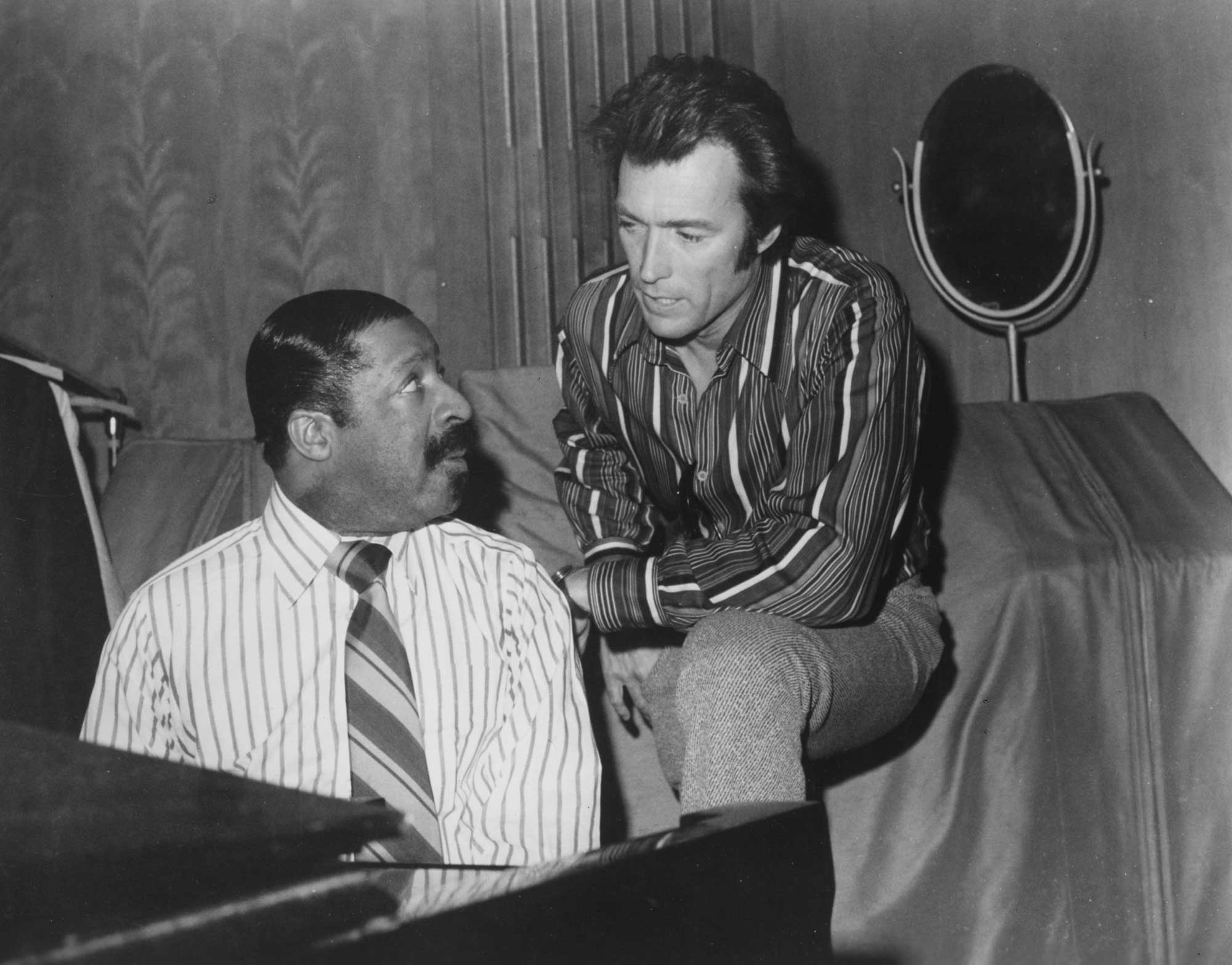 Black and white photo of two men, one seated at a piano wearing a striped shirt and tie, the other leaning on the piano wearing a striped shirt and tweed pants, engaged in conversation.