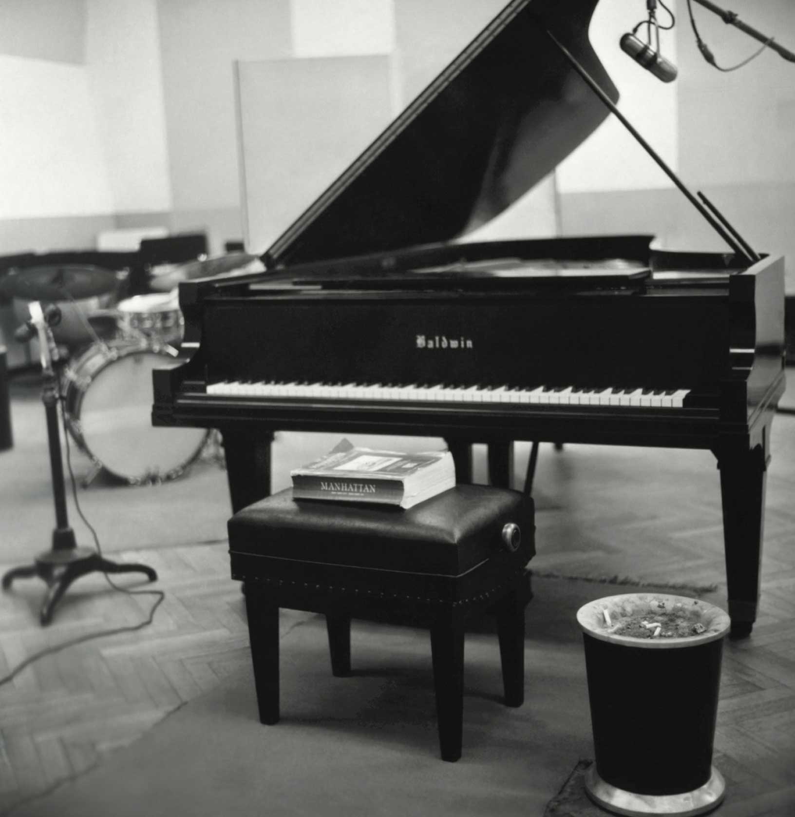Black Baldwin grand piano with an open lid in a music studio, a book titled 'Manhattan' on the piano bench, a drum set in the background, and a trash bin filled with cigarette butts nearby.