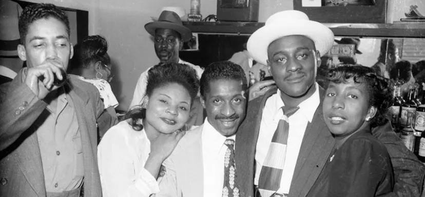 Black and white photo of five adults at a bar, three men and two women, smiling and posing closely together.