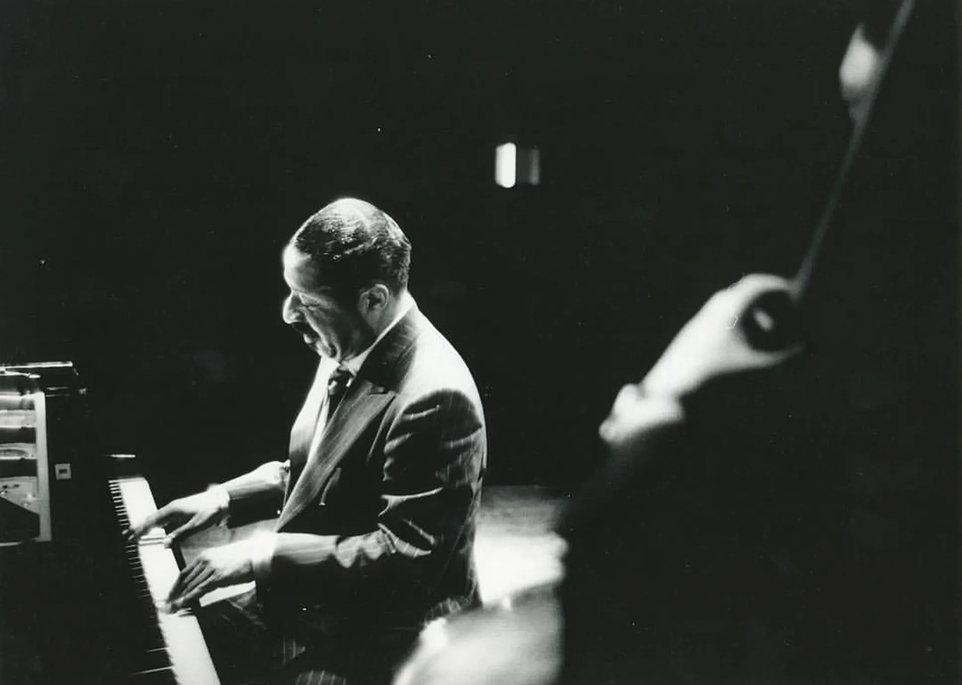 Black and white photo of Erroll Garner in a suit playing piano passionately on stage, with a blurred figure in the foreground holding a double bass.