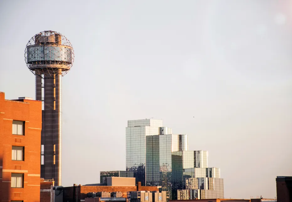 Urban skyline featuring the Reunion Tower with its geodesic dome and nearby modern glass buildings at sunset.