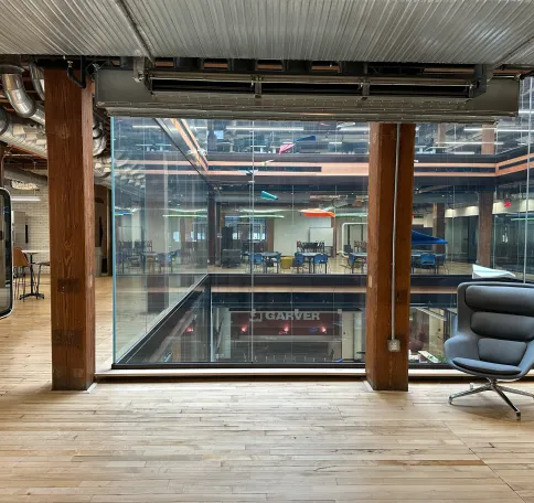Modern office interior with wooden floors, large glass windows, and a gray lounge chair on the right.