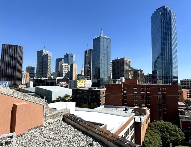 Downtown city skyline with tall modern buildings under a clear blue sky, viewed from a rooftop.
