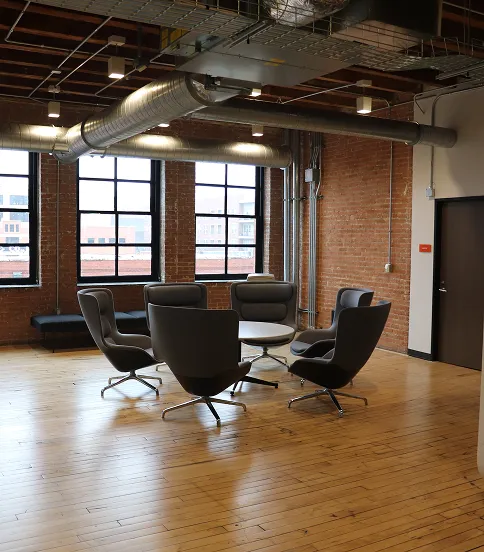 Modern office lounge with five gray swivel chairs arranged around a round table on a wooden floor.