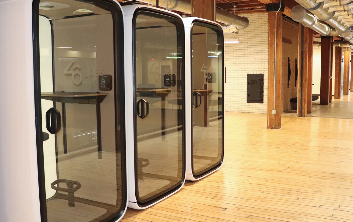 Three modern white phone booths with glass doors in a spacious office with wooden floors and exposed brick walls.