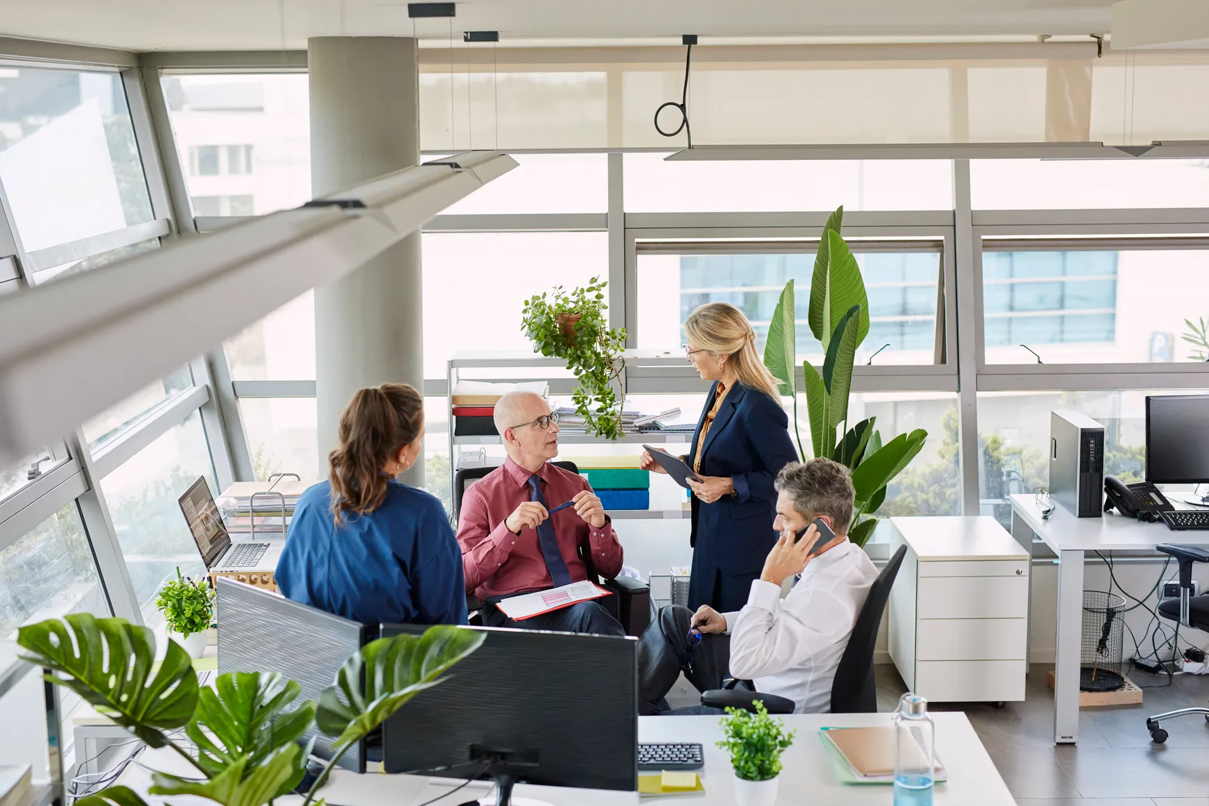Four business colleagues having a discussion in a bright modern office with large windows and green plants.