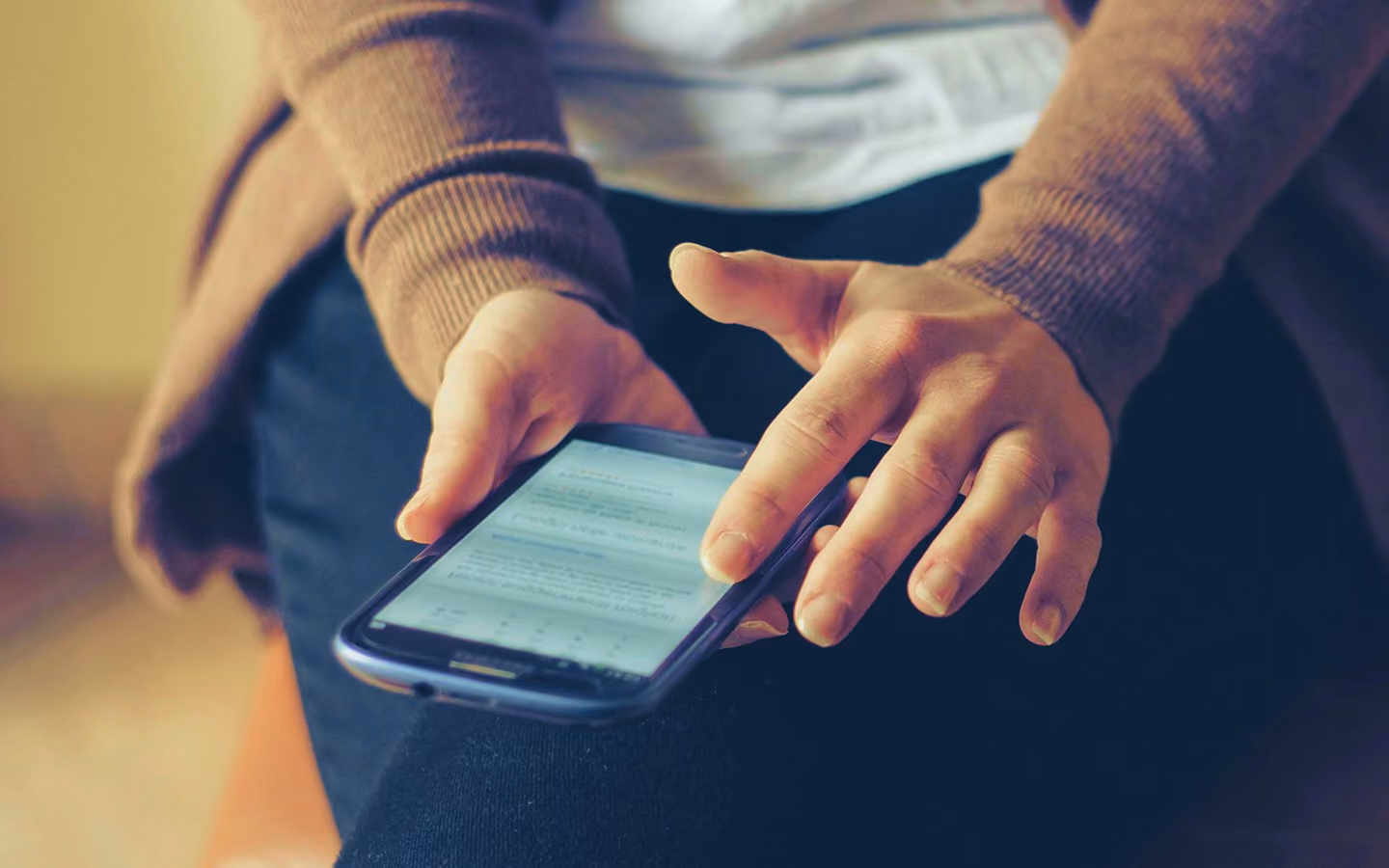 Person wearing brown sweater scrolling on a smartphone while sitting with crossed legs.