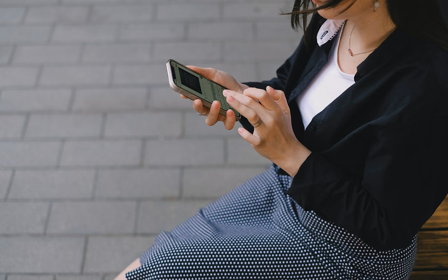Woman sitting on bench holding smartphone and typing with both hands outdoors on tiled ground.