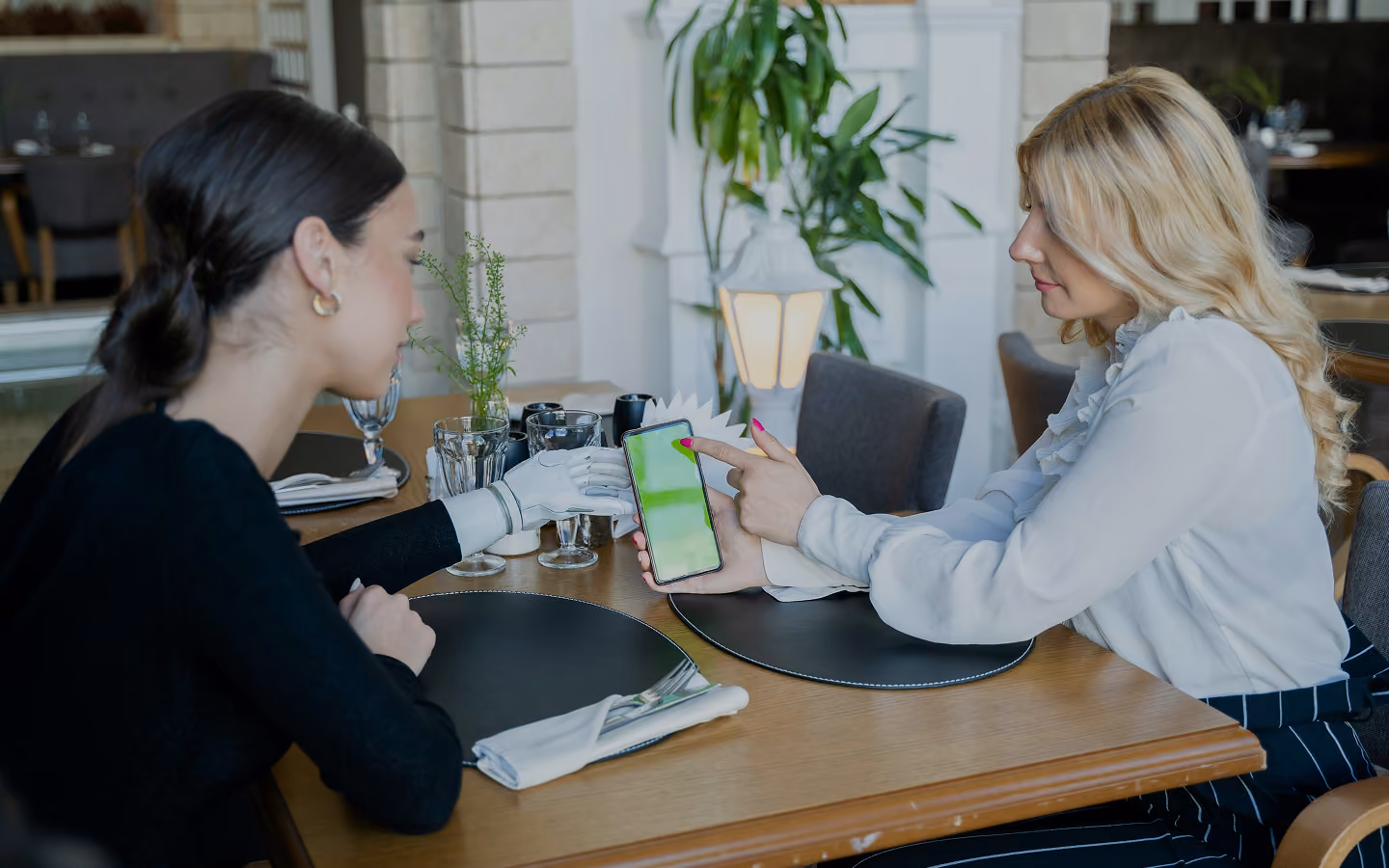 Two women sit at a table, one with a robotic arm, looking at a smartphone with a green screen.