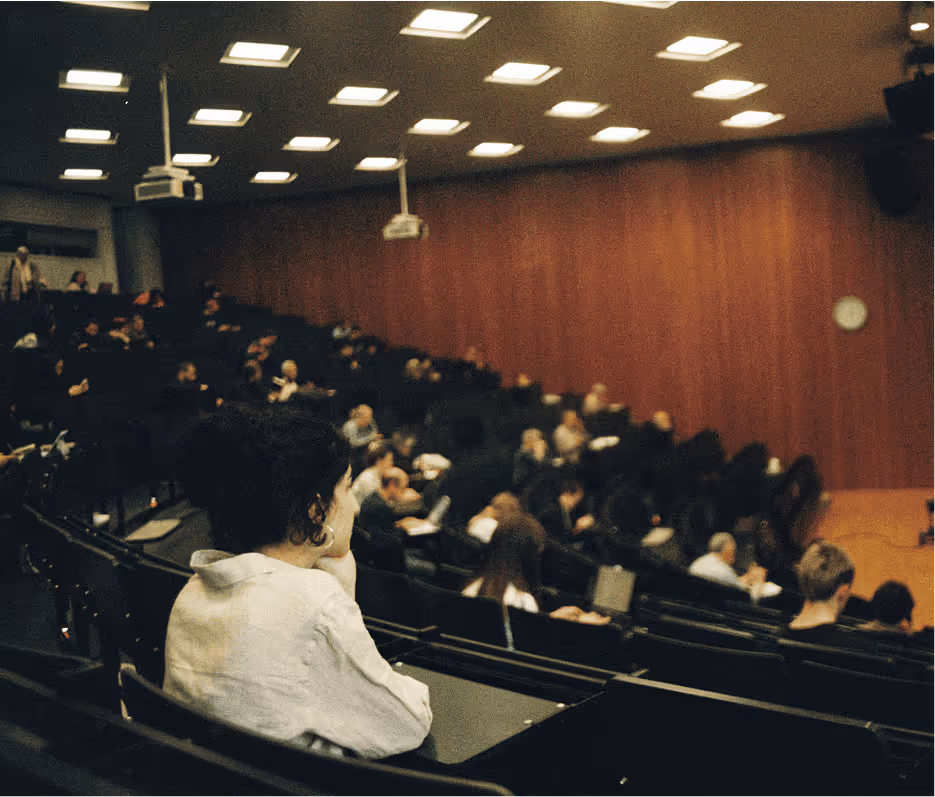 A person in a white hoodie sitting in a lecture hall auditorium, viewed from behind, with rows of seated audience members in the background under warm lighting.