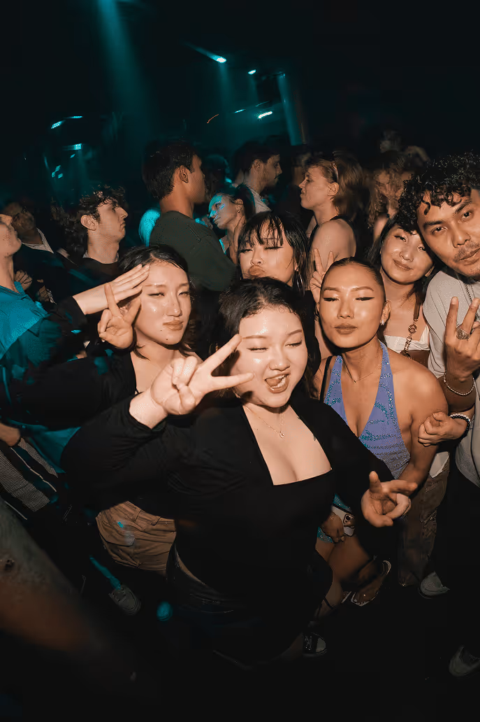 Group of young people at a party or club making peace signs and smiling at camera, with colorful neon lighting and crowded dance floor atmosphere.