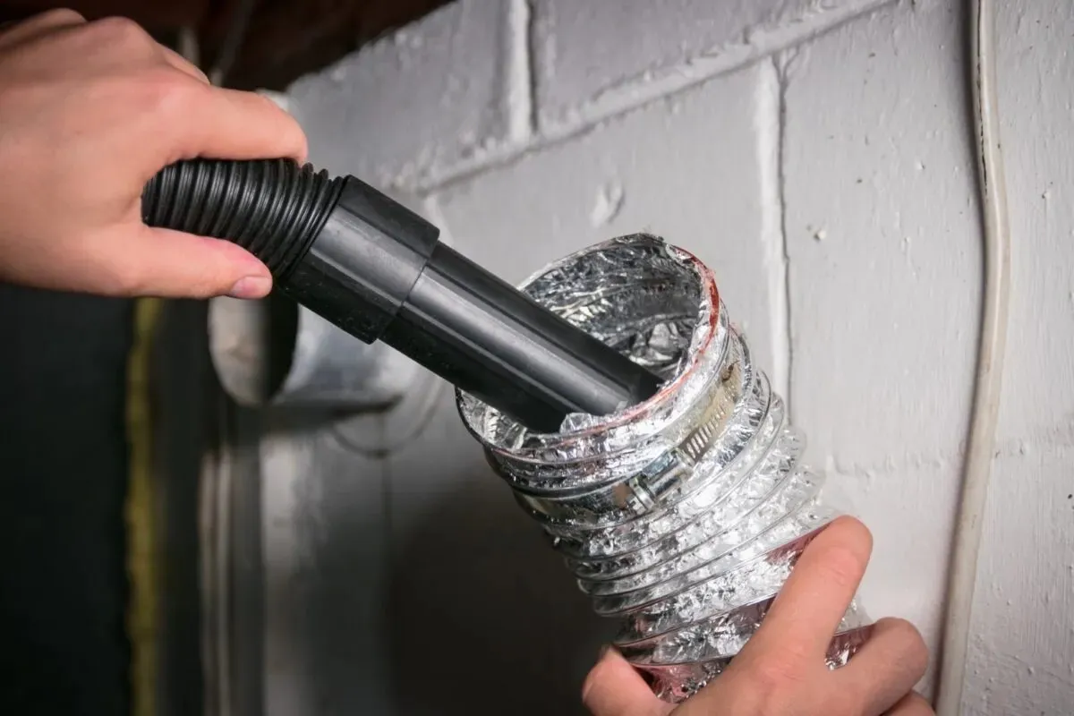 A person's hands are shown inserting a vacuum hose into a flexible silver dryer vent