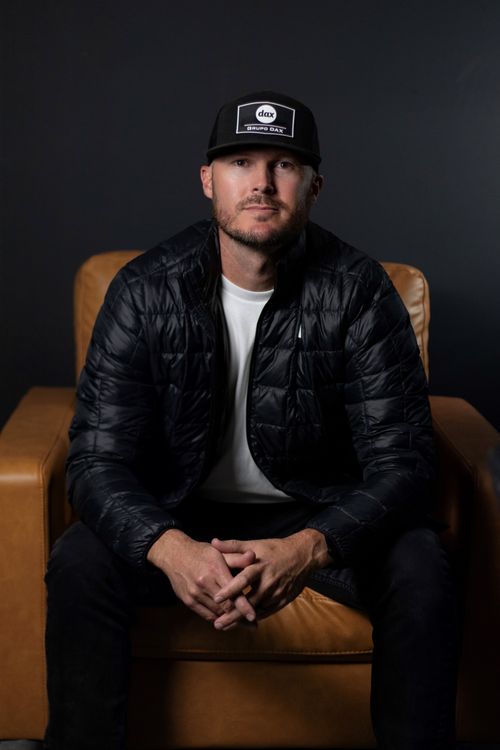 Un hombre caucasico sentado en un sillón cafe, en frente de una pared negra. El hombre esta inclinado un poco y recarga sus codos en sus piernas. Tiene una gorra negra en su cabeza que dice "Grupo Dax."
Su cara es seria pero agradable. Tiene barba y bigote muy cortos.