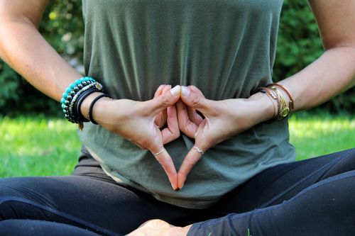 Close-up of Gabriella Ruocco seated in meditation pose, forming a grounding mudra with her hands resting over her abdomen, surrounded by nature