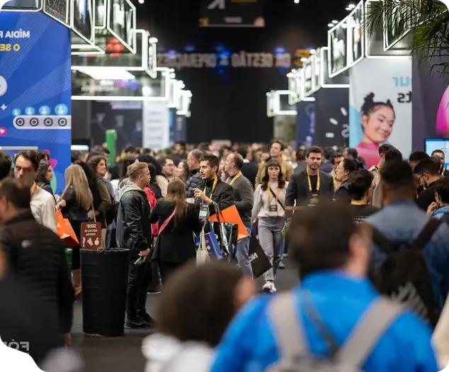 Multidão de pessoas caminhando e conversando em corredor de feira ou convenção com estandes iluminados.