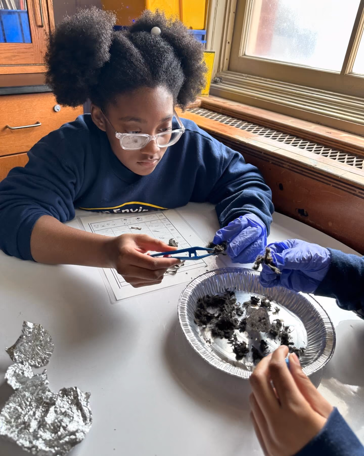 Student wearing safety goggles examines soil sample with tweezers during science lab
