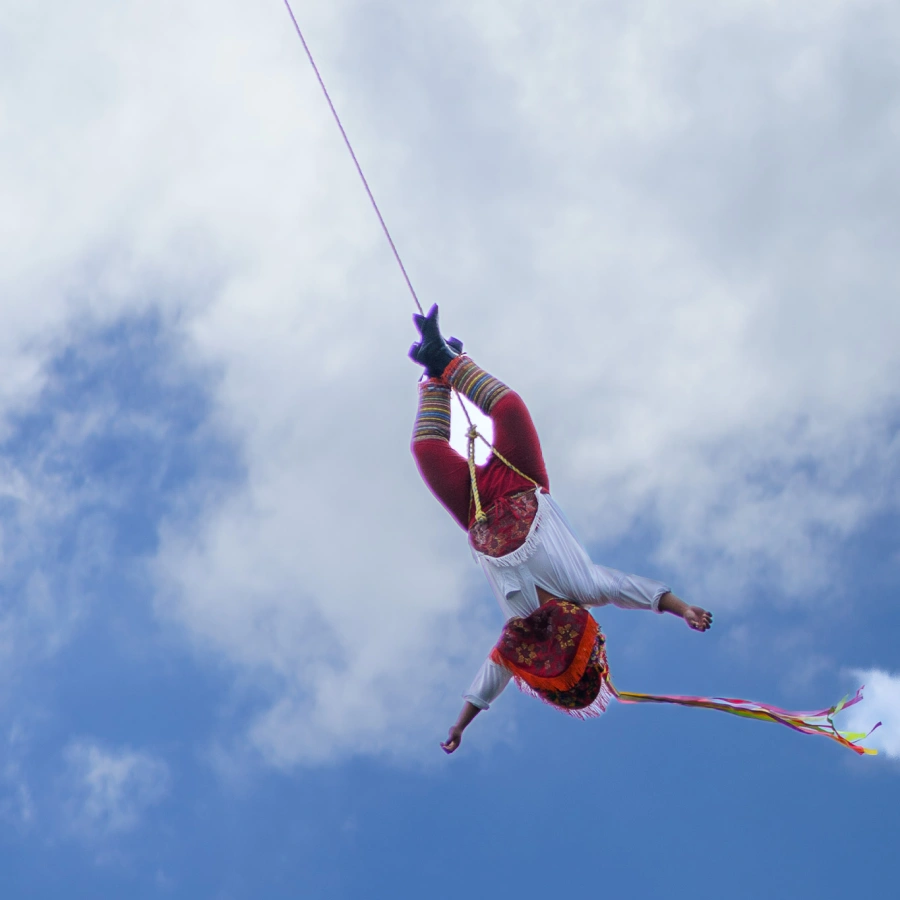 One man, wearing the colorful traditional costume of a Volador de Papantla, hangs upside down from a rope attached to the top of a tall pole, as part of a ceremonial aerial ritual in Mexico City