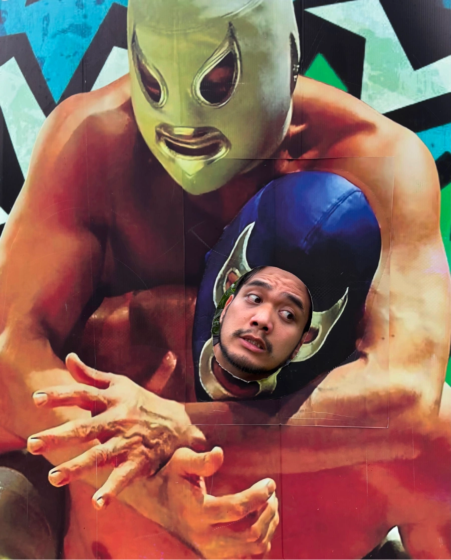 Dexter, our team member, poses inside a life-size luchador cutout, with the cutout’s arms framing his face.