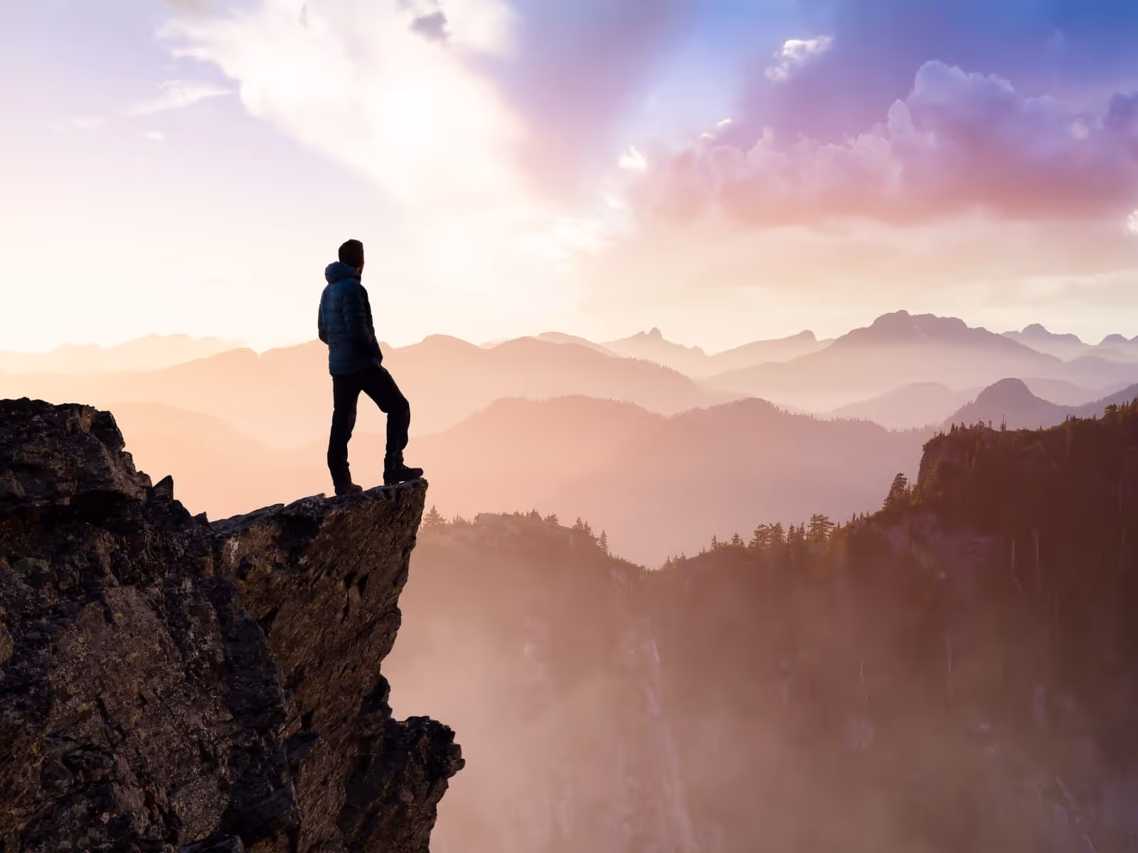 Man standing on top of mountain, at the peak. looking ahead at other mountains.