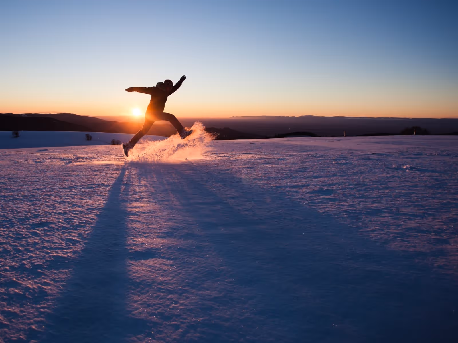 man jumping. against light. sunrise. snowy ground. snow being kicked in the air.