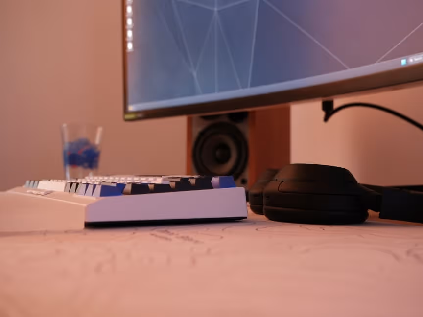 White keyboard with transparent wrist rest, black headphones, a monitor and speakers behind it.